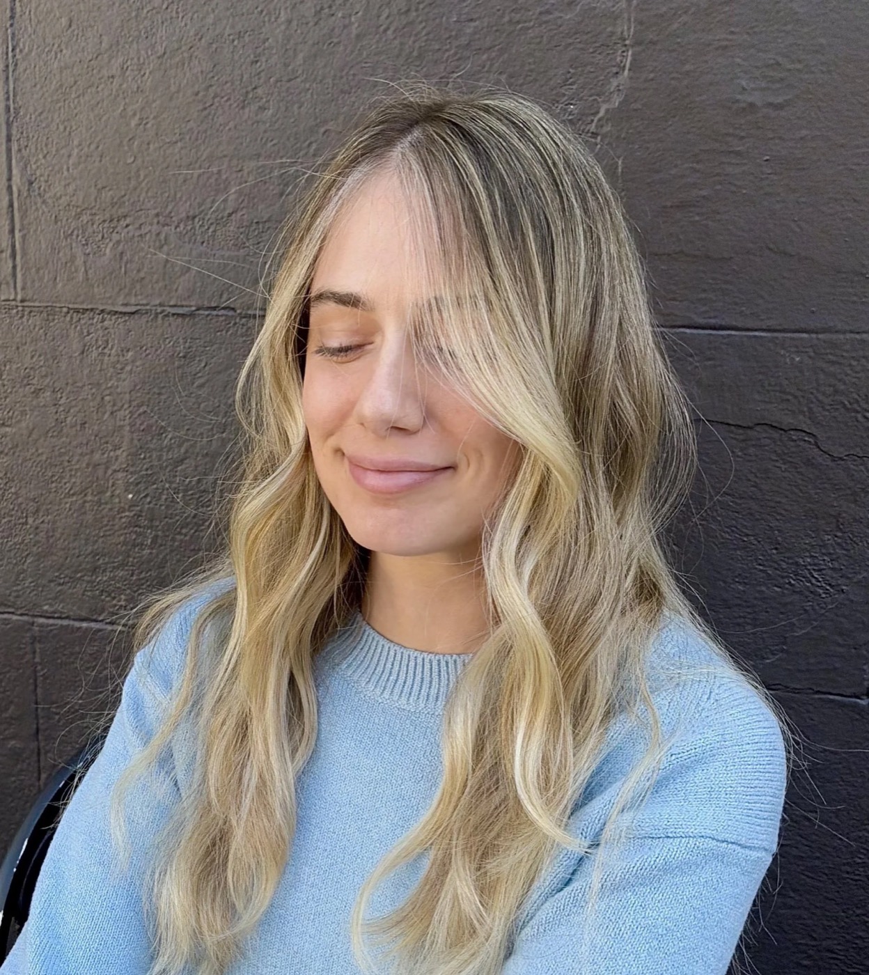 Close-up of a smiling woman with long blonde hair, wearing a light blue sweater, standing against a dark brick wall.